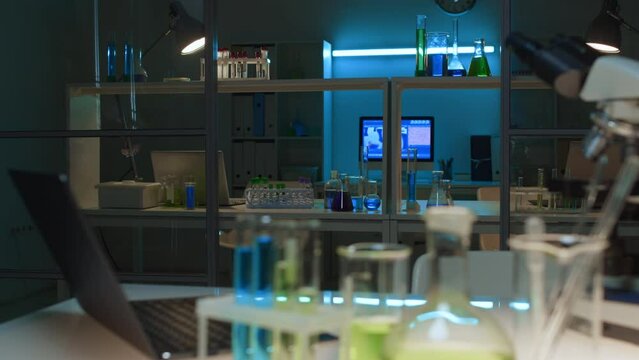 Close-up shot of worktop of scientist at chemical laboratory in dim light at end of workday, with laptop computer, microscope, test tubes, flasks, vials with bright green and blue solution, pipettes