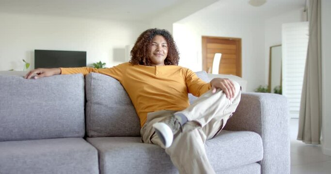 Portrait of happy biracial man with long hair sitting on sofa smiling in living room, slow motion