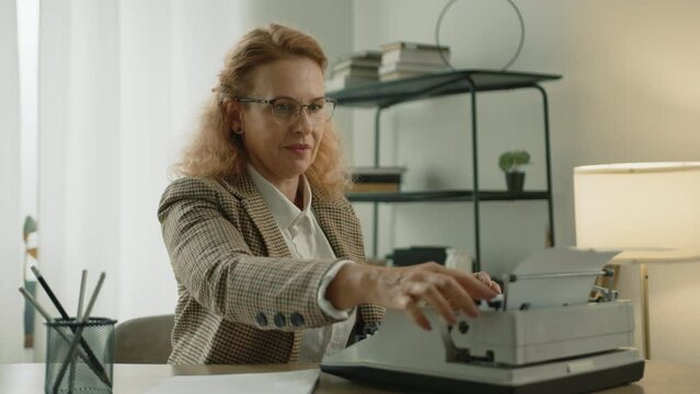 Enthusiastic mature woman in white T-shirt and checkered jacket focused on typing on old mechanical machine sitting at desk at home in cozy light room. High quality 4k footage