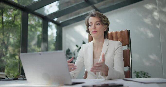 Young caucasian woman wearing glasses speaking at online conference or making a video call on a laptop in bright modern design business office space large sunlight windows and garden view