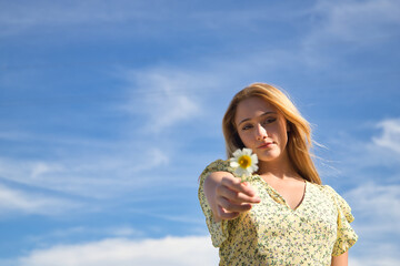 Young woman, beautiful and blonde in a yellow dress, offering a daisy to the camera, in the middle...