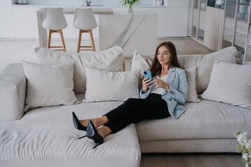 Relaxed young businesswoman laying on cozy couch at apartment holds phone thoughtfully looks away...