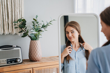 Pretty girl with beautiful healthy hair standing at mirror looks at herself smiles, preparing for...