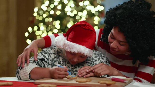 Middle-aged mother helping 10-year old sun to paint gingerbread cookie in blue color on kitchen board. Blurred background of Illuminated Christmas tree. High quality 4k footage