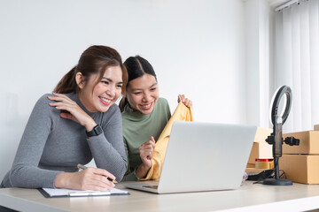 Young Asian female friends talk to a live camera on the phone to sell clothes together at home. and display clothing to customers in online sales