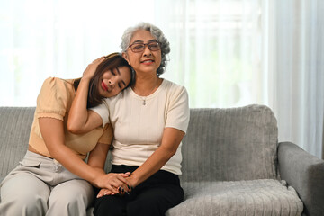 Beautiful grey haired elderly woman embracing her grownup daughter sitting on sofa at home