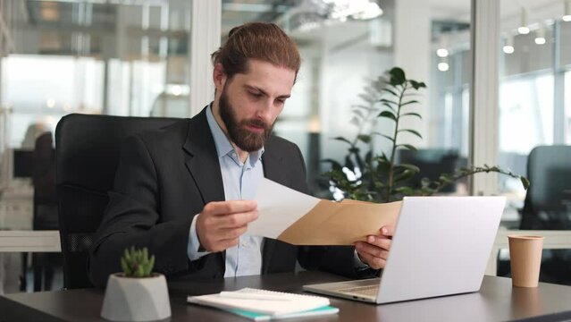 Excited young caucasian man working on wireless computer and feeling euphoric while reading paper letter correspondence. Overjoyed bearded male worker having pleasant news in postal envelope.
