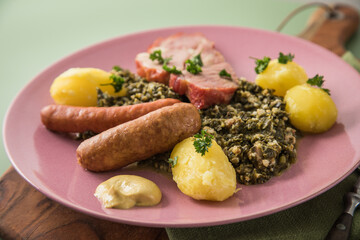 Oldenburger Grünkohl mit geräucherter Kohlwurst, Pinkel, Kassler Nacken, Kartoffeln, Senf, Petersilie auf Holzbrett, Serviette, Teller pink, und grün Hintergrund