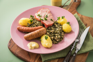Oldenburger Grünkohl mit geräucherter Kohlwurst, Pinkel, Kassler Nacken, Kartoffeln, Senf, Petersilie auf Holzbrett, Serviette, Teller pink, und grün Hintergrund