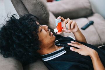 Man sitting on couch at home holding chest having difficulties breathing and using asthma inhaler to prevent shortness of breath   - obrazy, fototapety, plakaty