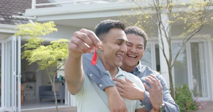 Portrait of happy diverse gay male couple with new house keys embracing in garden, slow motion