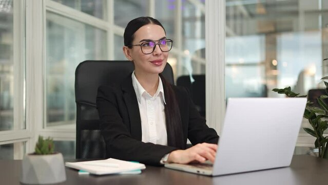 Positive female entrepreneur sitting and typing on laptop keyboard while dreaming about successful career. Caucasian brunette showing confident and good expectations in career ladder in office room.