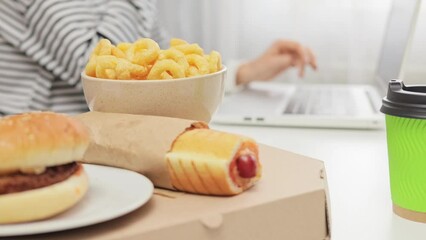 Closeup of unhealthy food hot dog onion snacks and burger and unknown woman working on laptop computer freelancer with favorite meals for dinner at work.