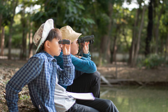Asian boys sitting by the pond and holding binoculars to watch birds and learning nature and environment together.
