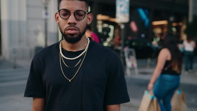 Portrait of young African American man stands confidently in bustling city street against backdrop of diverse pedestrians in a shopping district.Dressed in plain black t-shirt, layered gold necklaces