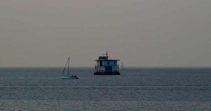 Two sailing boats float on the surface of the water at sunset, a lighthouse in the middle of the water