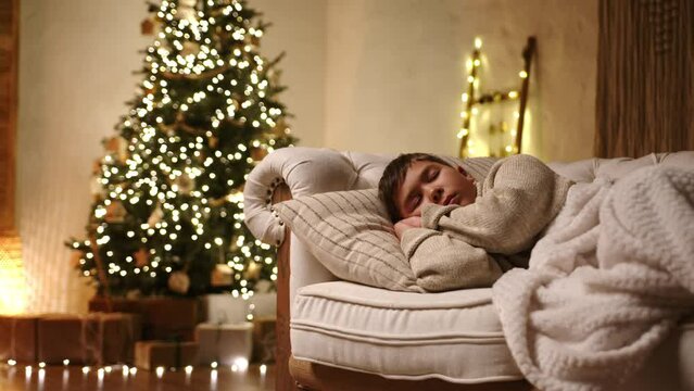 Young boy sleeping on modern light sofa, covered with plaid of milky color in a room with muffled light, plenty of gift-wrapped boxes under xmas tree. High quality 4k footage