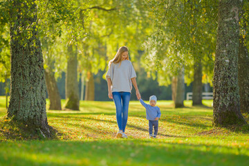 Young adult mother and baby boy walking on green grass through tree alley at park. Spending time together in beautiful autumn day. Front view. Lovely emotional moment. Peaceful atmosphere in nature.