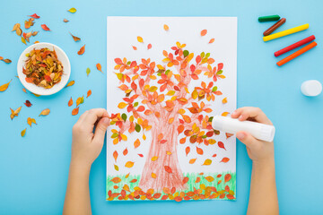Little child hands gluing and creating tree shape from colorful leaves on white paper. Light blue...