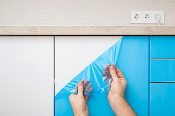 Young adult man hands removing blue protective polythene film from new kitchen cabinet doors under table top. Closeup. Front view. - obrazy, fototapety, plakaty