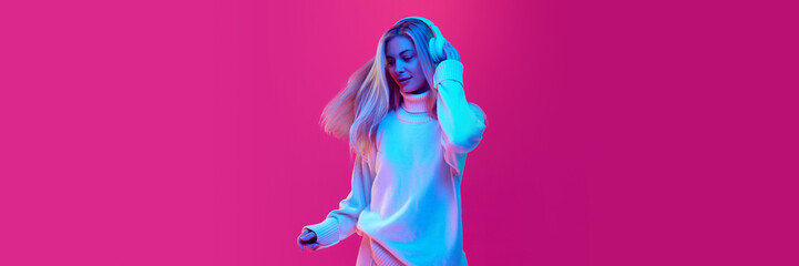 Banner. Attractive woman, student dancing while listening music in headphones against pink studio background in neon light, filter.