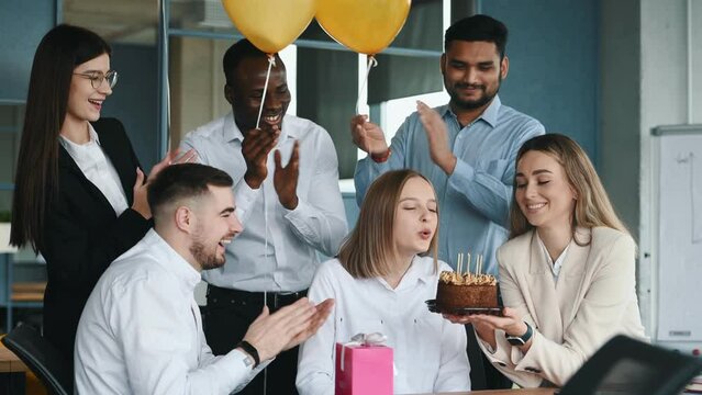 Young woman is working in office then getting birthday cake from colleagues making wish blowing candle enjoying surprise having fun with creative team