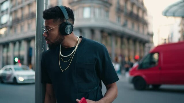 Pensive man with headphones in city, holding phone, urban soundscape and technology blend. Stylish millennial male enjoy listening to music in earphones in evening street outdoors. Slow motion