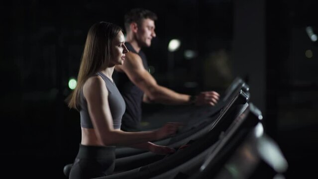 Wellness, young woman and man athlete walks and starts the run on a treadmill, two runners perform aerobic exercise and endurance training in the gym, nightlife.