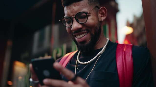 Smiling African American millennial man using smartphone on city street, concept of urban connectivity and technology. Male tourist enjoy online text messaging in roaming while travel with 5G