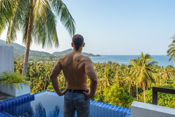 Young adult man standing on luxury tropical pool villa resort on balcony and looking forward on sea...