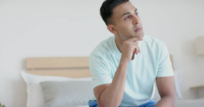 Contemplative biracial man sitting on bed in sunny bedroom thinking, slow motion