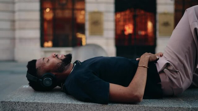 Male technology user enjoying perfect playlist that uplifts the mood and brings sense of pleasure while lies back with closed eyes in urban setting, Black man enjoying online music beat with app