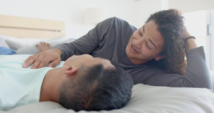 Happy diverse gay male couple lying on bed talking at home, slow motion