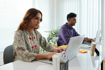 Serious young Indian woman working on laptop in office, answering email from clients