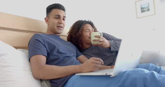 Happy diverse gay male couple on bed, having coffee and using laptop in the morning, slow motion
