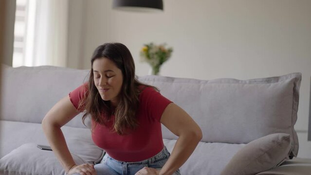 Caucasian woman standing up from a couch at home experiences sudden back pain. Young adult female suffers from pain in her back raising from sofa. Girl rubbing back suffering from cramps in lumbar
