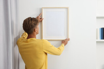 Young man hanging picture frame on white wall indoors, back view