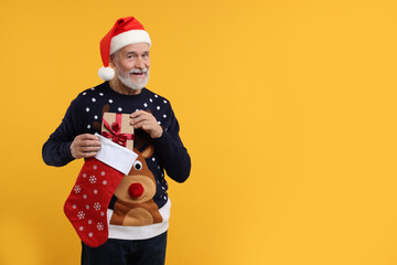 Senior man in Christmas sweater and Santa hat taking gift from stocking on orange background. Space for text