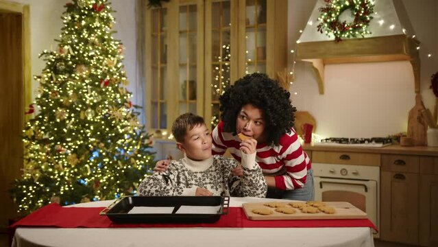 Happy mum and minor son in festively decorated kitchen with xmas tree with garland, baking gingerbread men, taking a sample of homemade bakery, hugging. High quality 4k footage