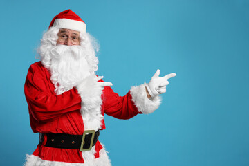 Fototapeta na wymiar Merry Christmas. Santa Claus pointing at something on light blue background, space for text