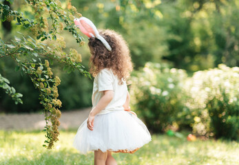 Easter egg hunt. Girl child Wearing Bunny Ears Running To Pick Up Egg In Garden. Easter tradition. Baby with basket full of colorful eggs.