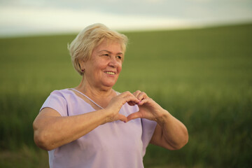 Cheerful elderly woman with a heart gesture amidst nature; warmth radiates. Reflects the connection between outdoor activity and senior wellness.