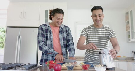 Happy diverse gay male couple preparing healthy fruit smoothie in kitchen, copy space, slow motion