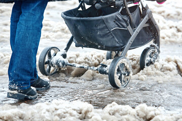 Woman push baby stroller through wet snow in winter season. Woman with baby carriage walk through...