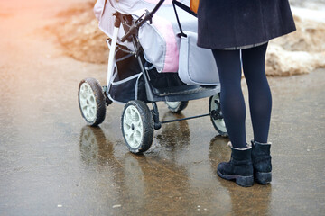 Trendy woman with baby carriage walk on wet sidewalk in the city in winter season. Fashionable...
