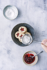 Eine Hand streut Puderzucker über Weihnachtskekse mit Erdbeerkonfitüre in einer Schüssel. Draufsicht.