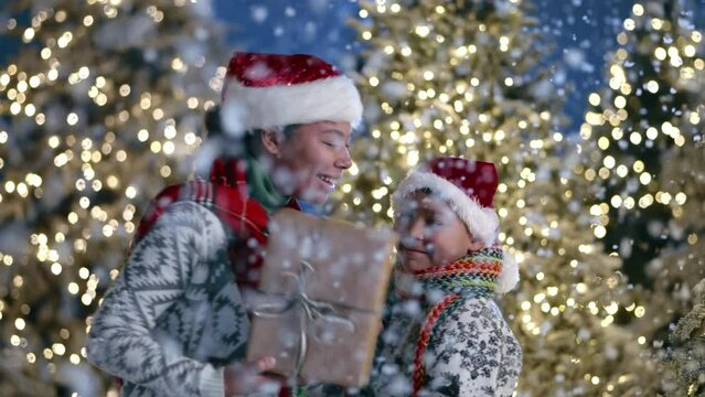 Happy mother, son in santa hats, sweaters with ornament hugging each other with fluffy snowflakes in foreground, illuminated Christmas trees on background. High quality 4k footage