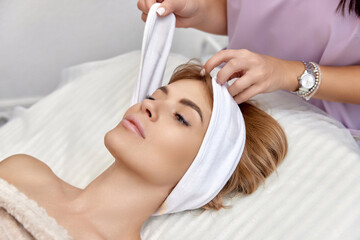 Obraz na płótnie Canvas Cosmetologist putting white headband towel on female patient head in beauty salon