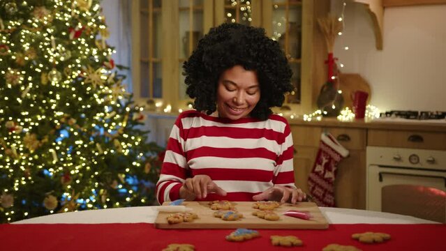 Smiling African American female coloring gingerbread cookies in a kitchen with modern furniture and garlands, illuminated xmas tree. Christmas spirit. High quality 4k footage