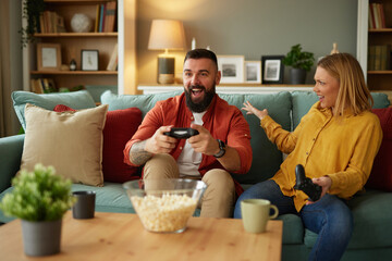 Couple sitting on couch with joysticks and playing video game in living room